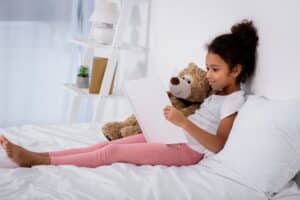 adorable african american kid reading book and hugging teddy bear on bed