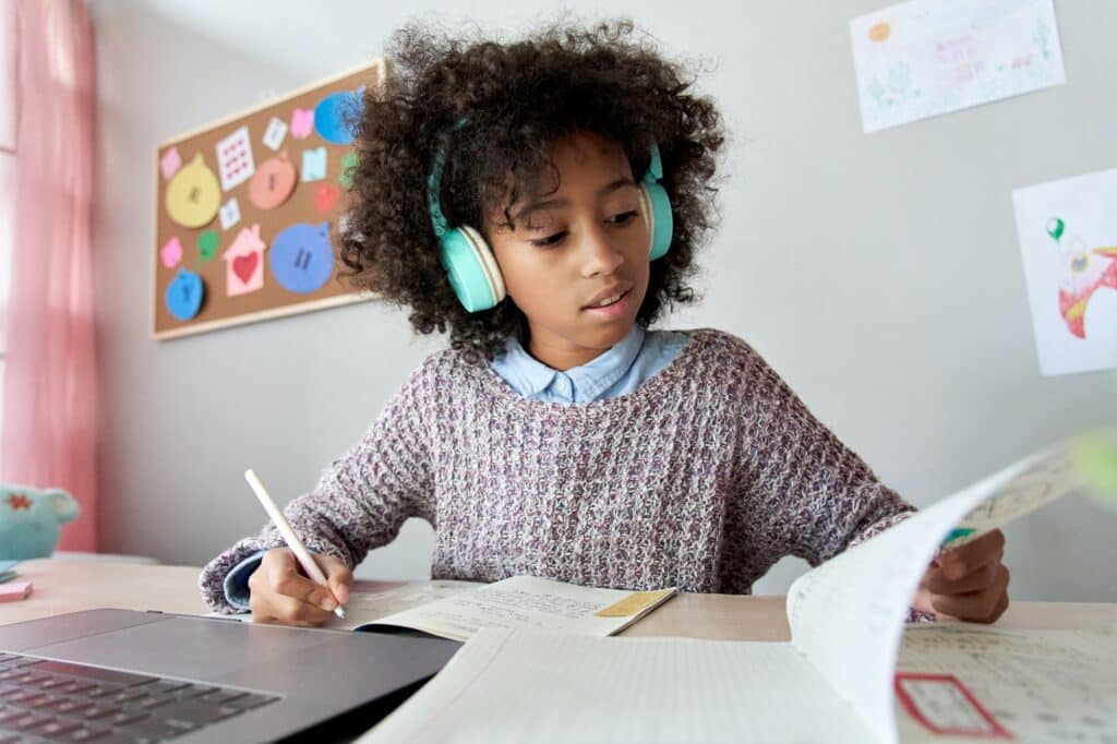 Smart african kid girl wears headphones learning online doing homework at home.