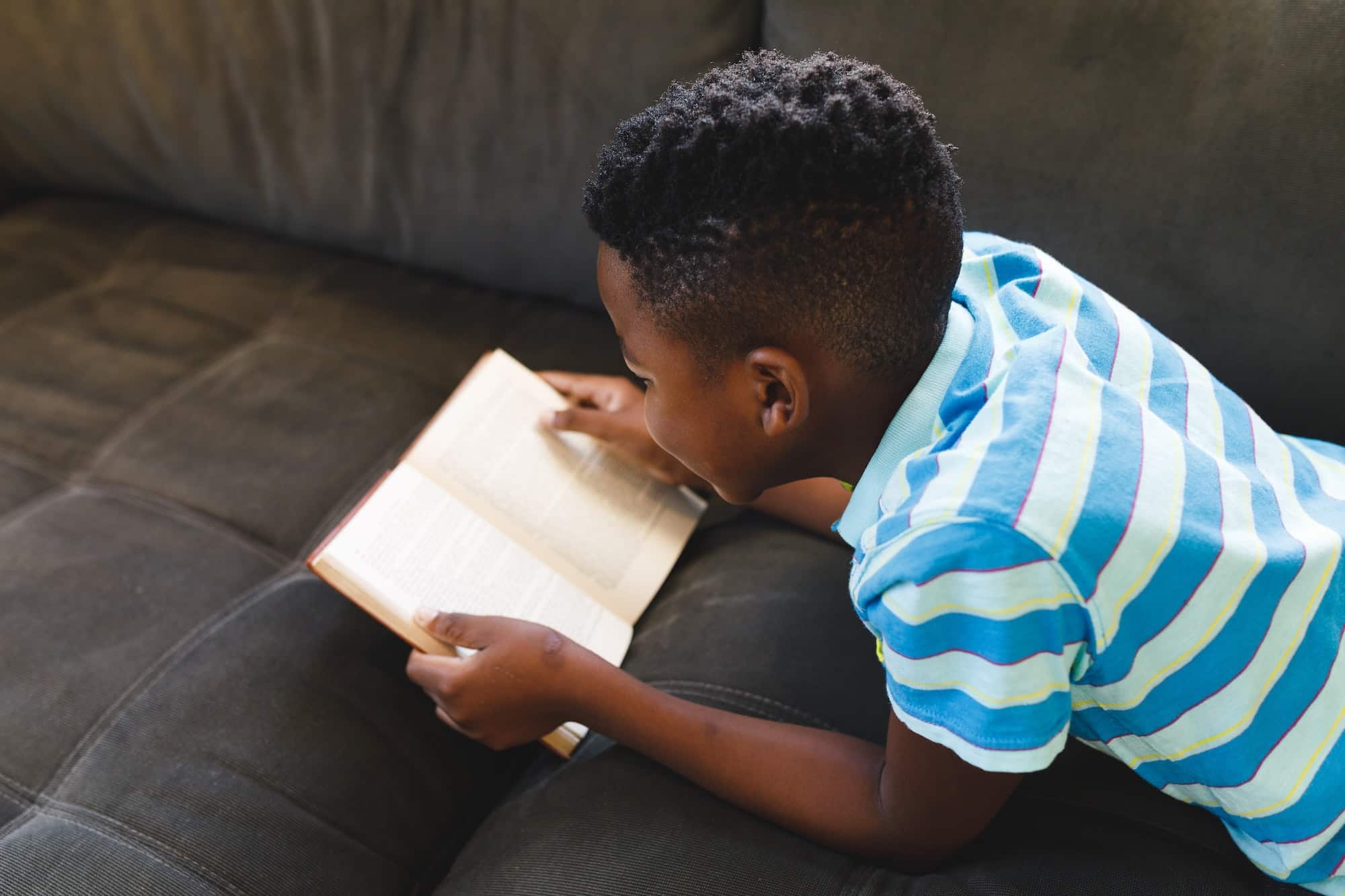 African american boy reading book and lying on couch in living room
